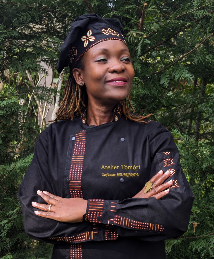 Taoficatou Hounkponou, en veste de cheffe noire aux motifs wax, souriante devant un feuillage
